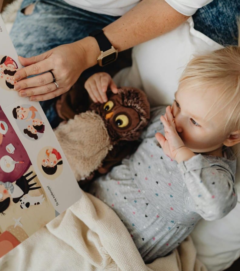 A young girl reads a storybook with her parent and a cuddly owl toy, sharing a cozy bonding moment.