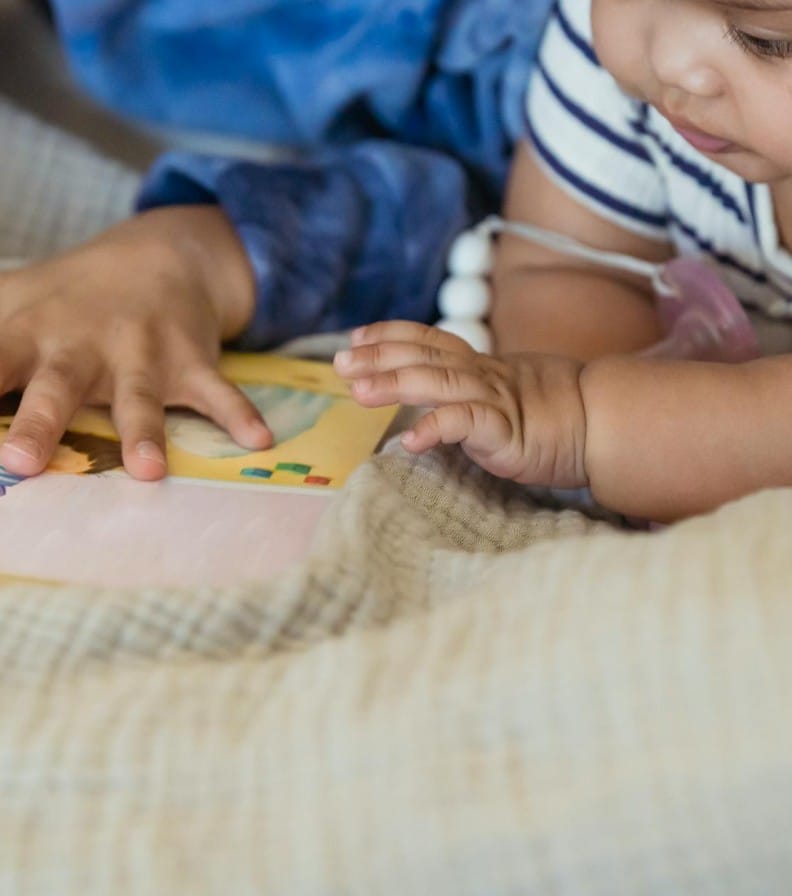 Unrecognizable boy reading interesting book while lying on blanket near cute little baby on sofa in cozy room at home