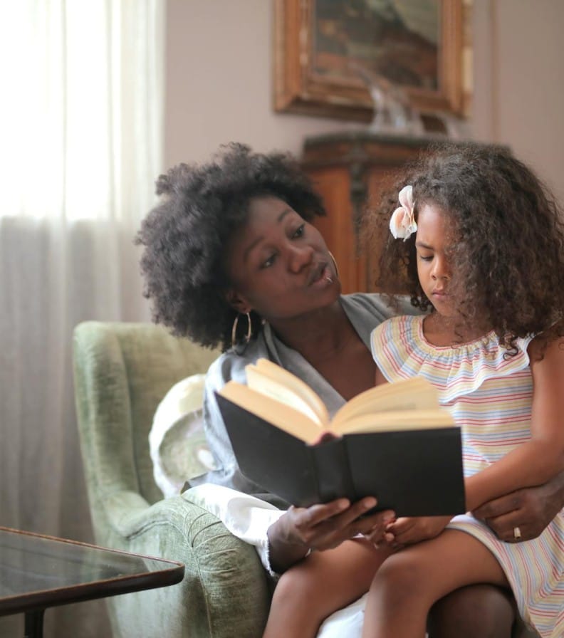 An African American mother reads a storybook to her daughter on a sofa, creating a warm bonding moment.