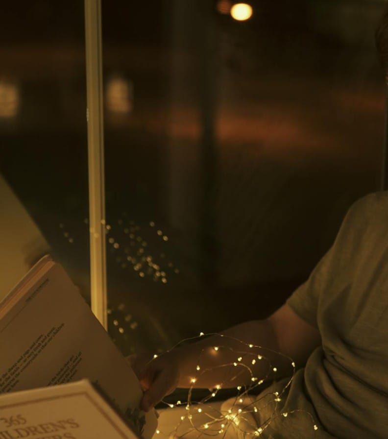 A young boy reads a children's storybook next to a window, illuminated by warm lights.