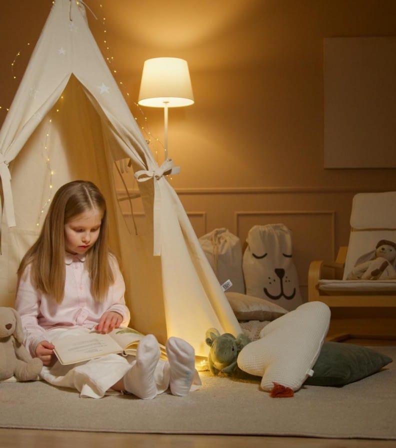 Child enjoying a book inside a cozy illuminated tent in a softly lit room.
