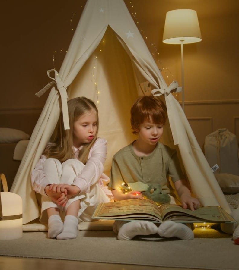 Two children enjoy reading in a cozy indoor playroom tent, surrounded by soft lighting.