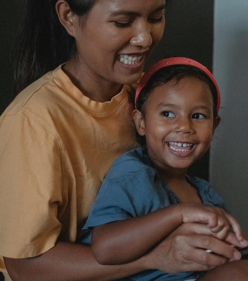 Smiling ethnic crop woman hugging playful girl while reading interesting story together near black wall during free time in apartment