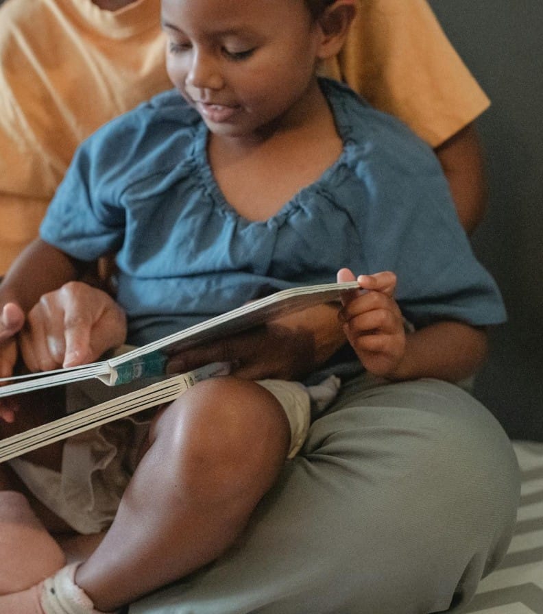 Unrecognizable ethnic mother hugging little girl and reading interesting book while sitting on couch near wall during weekend in apartment