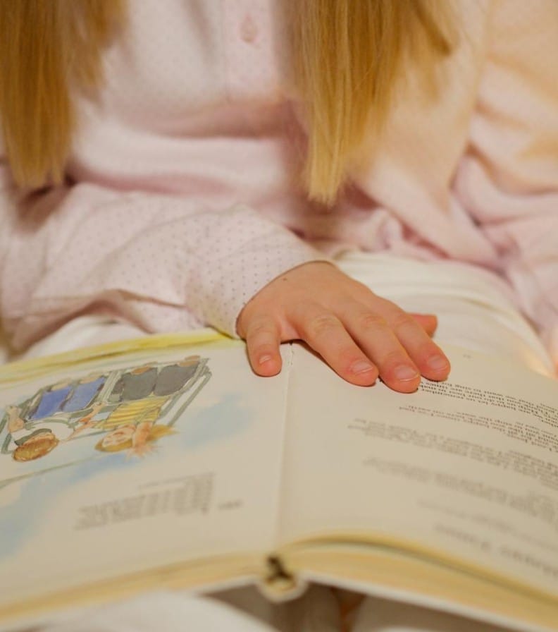 A young girl in pajamas reading a storybook in a warm, cozy bedroom setting.